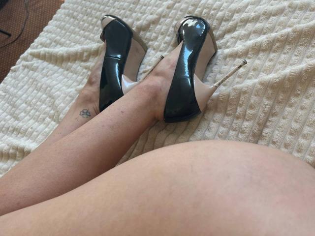 MoonLiza's legs in black shiny high heels, showing a small tattoo on her ankle, with her bare ass in the foreground. Her skin is pale, and the texture's a white knitted blanket.