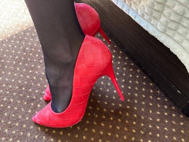 MoonLiza's shiny red high heels with a quilted pattern, standing on black pantyhose, on a brown carpeted floor, next to a dark wooden bed frame and white quilt.