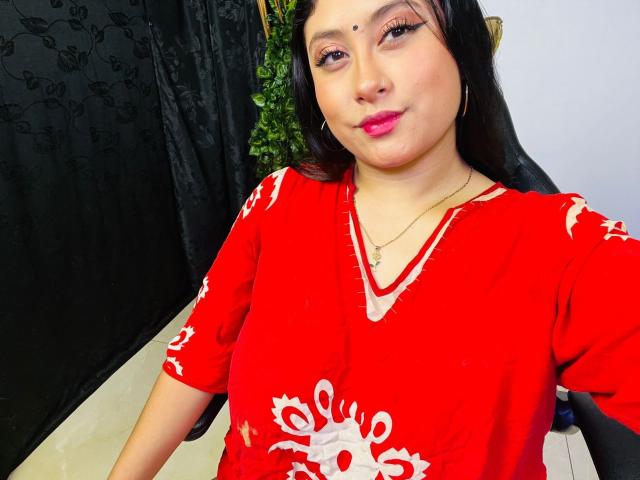 KalpanaAisha with medium tits, wearing a red dress with white floral patterns, has pink lipstick, black hair, and a black dot on her forehead, sitting in a chair.