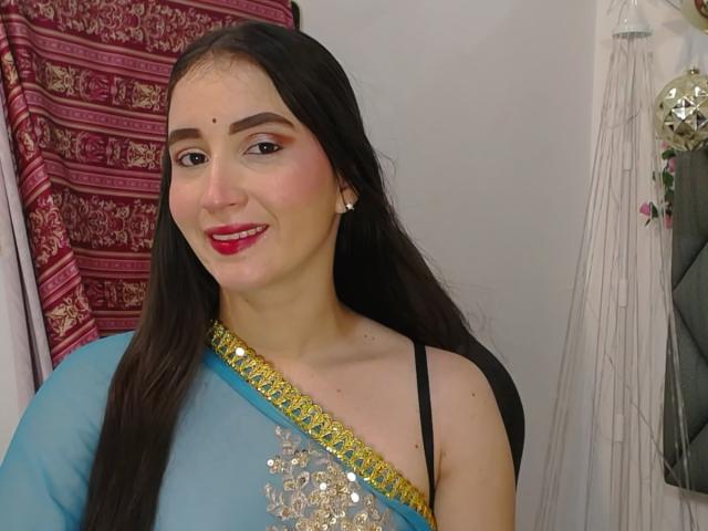 JalimaMona with long black hair, wearing a blue sari with gold and silver embroidery, black strap, red lipstick, and small earrings, smiling at the camera.