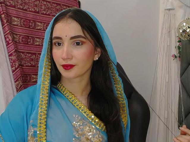 JalimaMona, wearing a blue burqa with gold and silver embroidery, has long black hair, red lipstick, and a mole above her right eyebrow. Her tits are covered by the burqa.