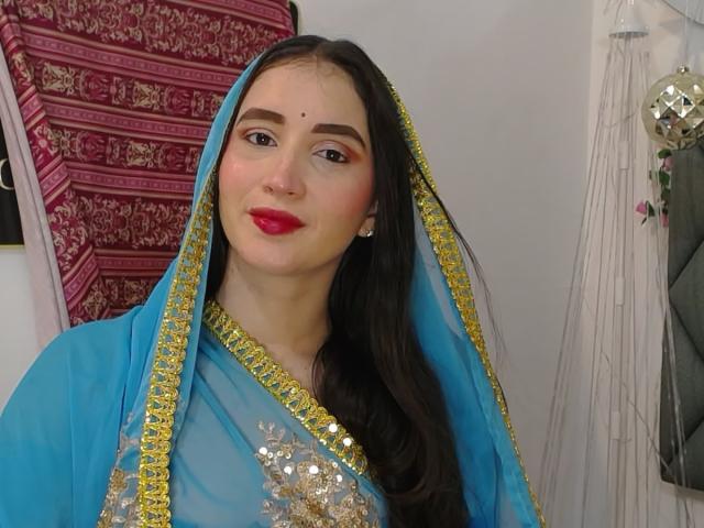 JalimaMona with long black hair, blue and gold dress, red lipstick, and a mole above her right eyebrow, looking at the camera, smiling. She's wearing a transparent veil.