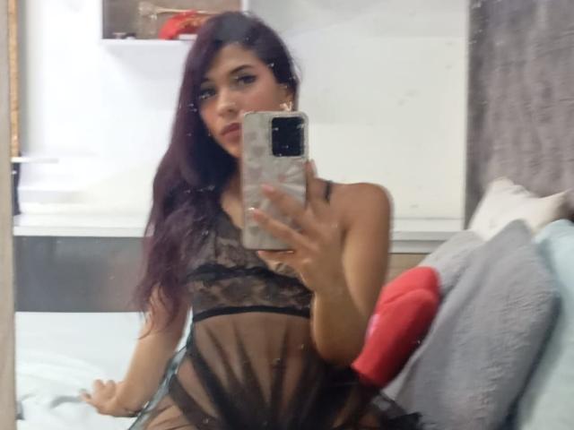 DanikaSerov with long purple hair, wearing a sheer black lace dress, sitting on a bed, taking a mirror selfie, showing her small tits and slim figure.