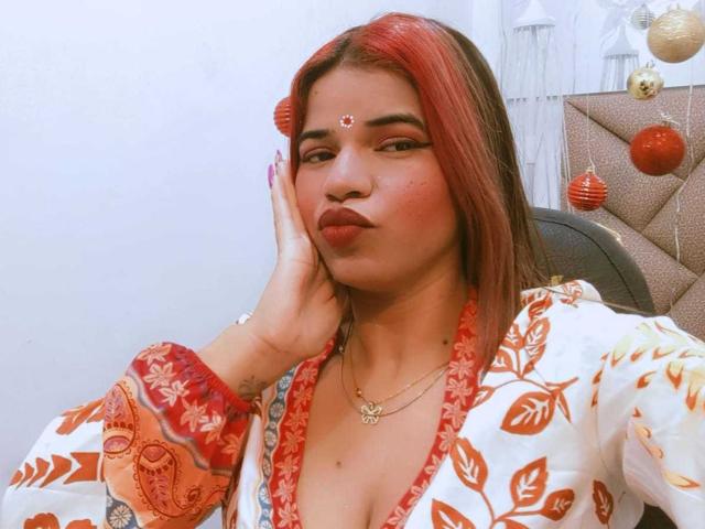 KairaDayamai with red-dyed hair, wearing a floral red and white top, has her hand on her cheek, showing her pouty ass and a red bindi on her forehead.