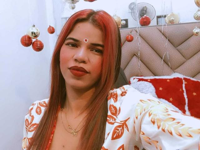 KairaDayamai with red hair, white floral robe, red lipstick, and a red dot on her forehead, sitting on a bed with red and white pillows. Boobs are not visible.