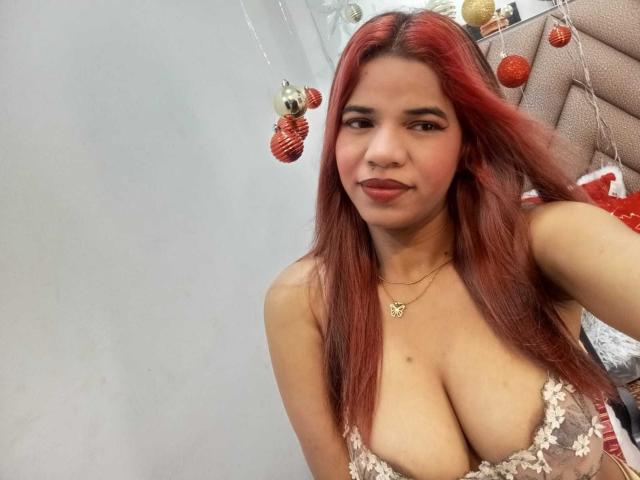 KairaDayamai with red hair, medium skin, and large tits in a sheer lace top, smiling at the camera, wearing a gold necklace with a heart pendant.