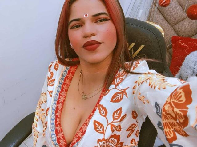 KairaDayamai with red hair, wearing a floral kimono, deep cleavage, red lipstick, and a forehead bindi, smiling, sitting in a black chair, wearing a necklace.