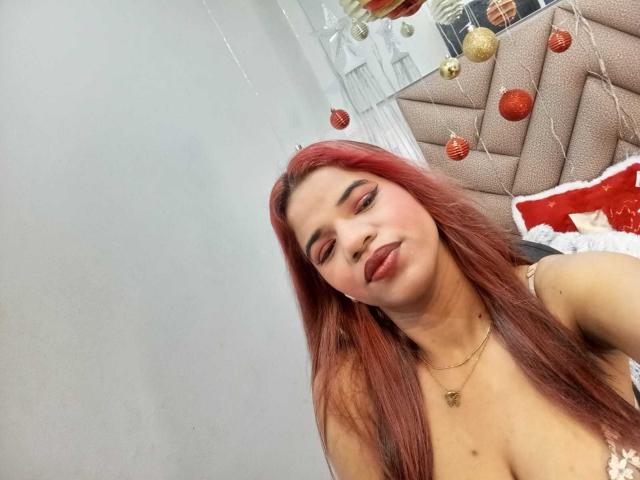 KairaDayamai with red hair, pink eyeshadow, and lipstick, wearing a gold necklace, showing her boobs, sitting in front of a beige, patterned headboard with red and gold ornaments.