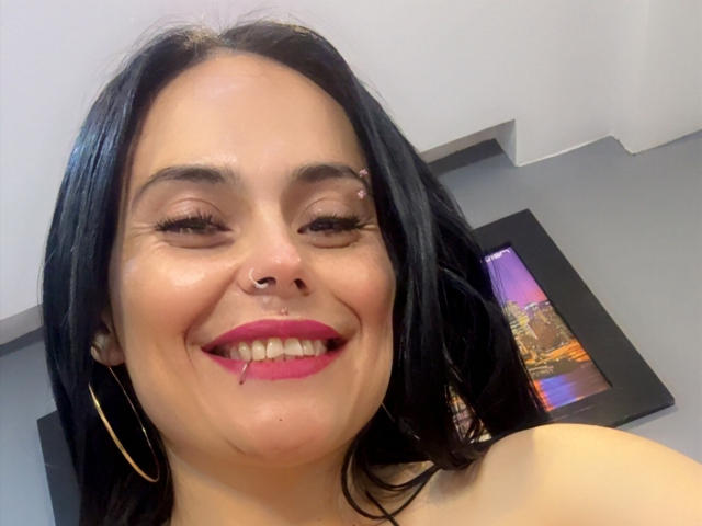 CharlotteCrofort with black hair, nose ring, and septum piercing, smiling with pink lipstick, hoop earrings, and a small cut on her lower lip. Her tits are slightly visible.