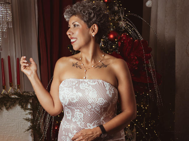 JuliaNewell stands in a Christmas-themed setting. She has short, curly gray hair and a tattoo of black vines on her chest. She wears a strapless white lace dress that reveals her medium-sized breasts. She's holding her right hand up, smiling to the side. Behind her is a decorated Christmas tree with red ornaments and a red bow, and a chandelier with red candles. Her skin is dark brown and she wears a black wristwatch. The image is warm and festive.