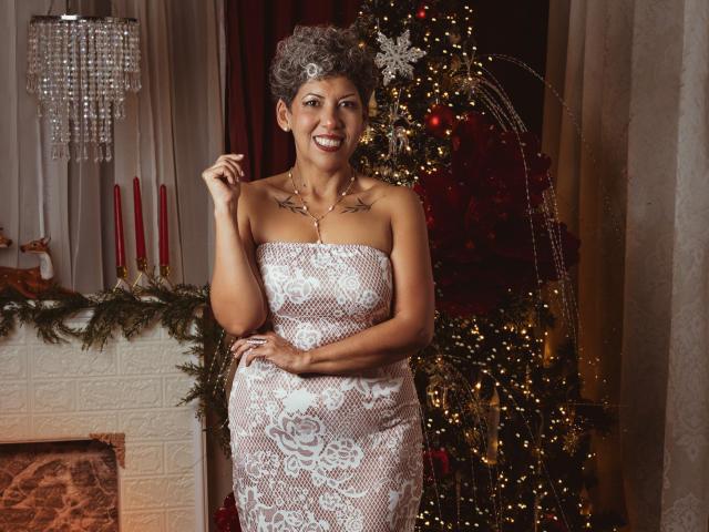 JuliaNewell stands in front of a Christmas tree, wearing a white, lace strapless dress. She has curly gray hair, smiling with visible teeth, and holding her right hand near her face. She has tattoos on her upper chest. The Christmas tree is decorated with red ornaments and white lights. Red candles are on the left, and a chandelier hangs above. The lace dress has floral patterns. The image has warm lighting.