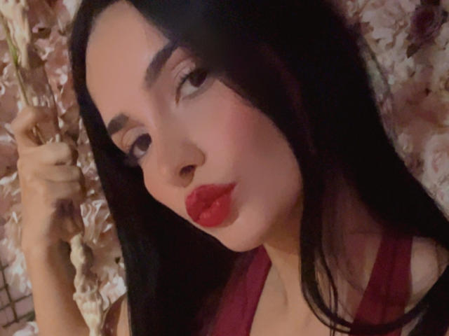 SexyWomanX with long black hair, red lipstick, and thick eyebrows, pouting with her hand in her hair, wearing a red top, looking seductive.