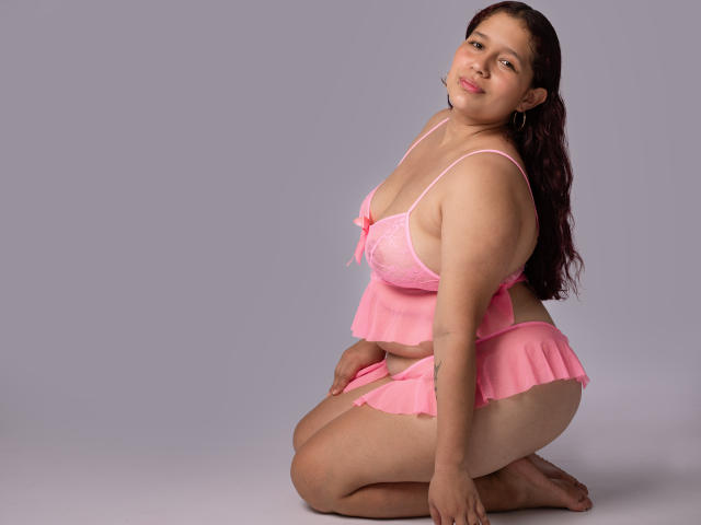 AliceRousee is a curvy woman with light brown skin, dark wavy hair, and pink lingerie. She's kneeling, showing her ass and small tits, with a confident smile.