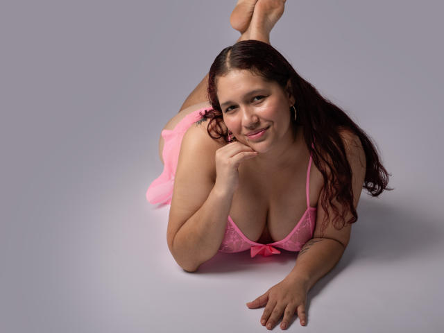 AliceRousee lying on her stomach, wearing pink lace lingerie, with her tits prominently displayed, legs up, smiling, brown wavy hair, and her hand near her mouth.