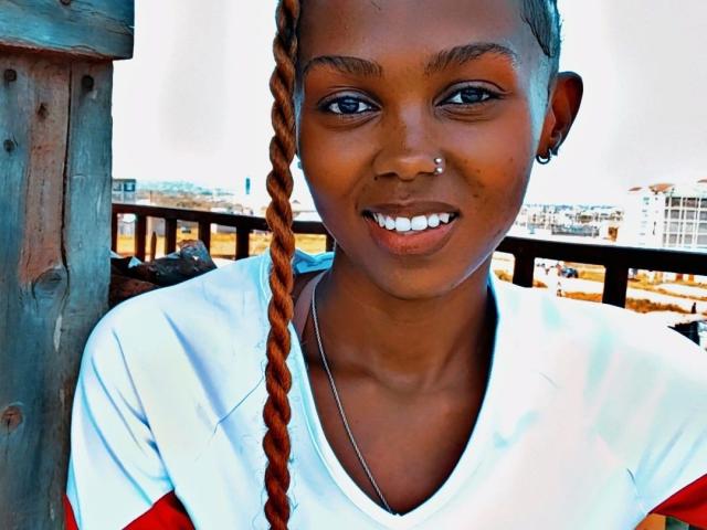 NastyVee-hot with dark skin, braided brown hair, nose piercing, and silver earring, smiling in a white and red shirt, standing next to a wooden structure.