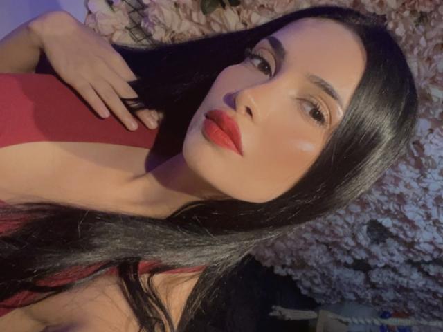 SexyWomanX with long black hair, red lipstick, and a red dress, her hand on her chest, looking at the camera, showing off her smooth, fair skin and sexy face.