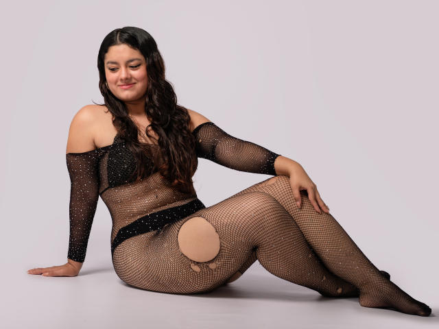 MimiRose with long dark hair, wearing a black, sheer, fishnet bodysuit with cutouts, sitting, smiling, showing her tits and pussy through the mesh, on a light gray floor.