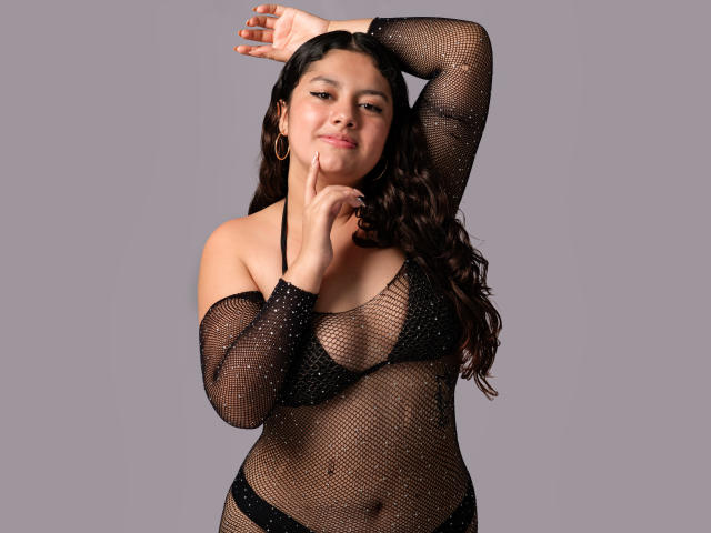MimiRose has long dark hair, wearing a black fishnet top with small studs, showing her tits and black bikini bottom, with one arm raised and finger on chin.