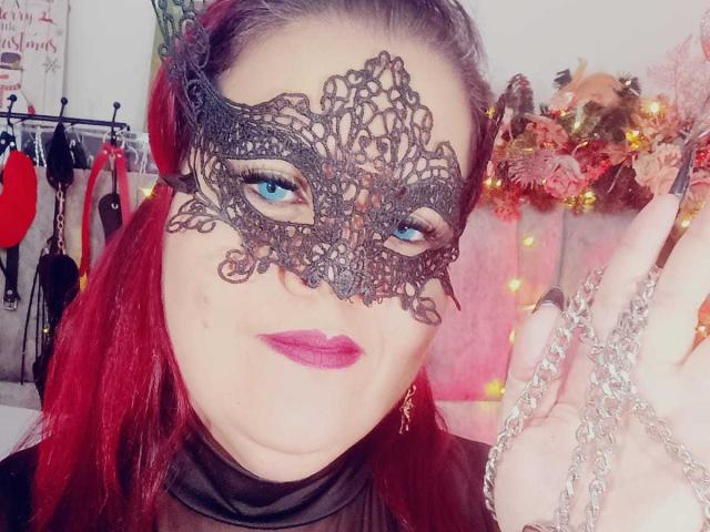 VanesaMoka with red hair, black lace mask, blue eyes, pink lipstick, holding a chain, showing off manicured nails, in a sexy, playful, and elegant pose.