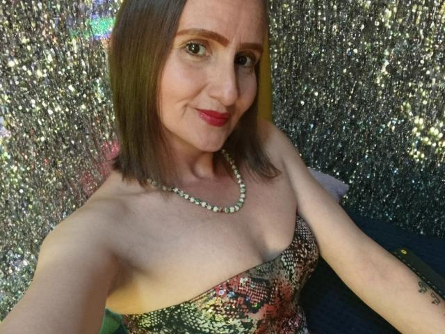 Rubi Anderson, con cabello castaño hasta los hombros, lápiz labial rojo, collar de perlas, vestido estampado sin tirantes, sonriendo, mostrando senos pequeños y un tatuaje en su brazo derecho, en un selfie..