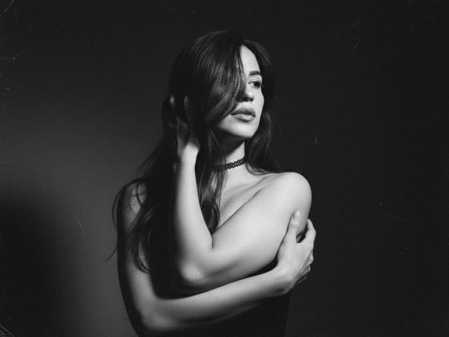 MelisaDaltonn is topless with long dark hair, hand in hair, other hand covering small breasts, choker necklace, black background.