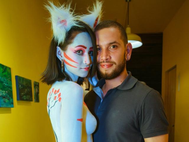 SpicyFran, con pintura blanca en la cara, rayas rojas y orejas de gato, junto a un hombre con barba que lleva un polo negro, ambos sonriendo, en un interior, bajo una luz colgante..