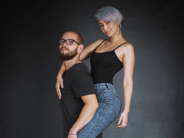 SpicyFran, con el pelo corto y morado, una camiseta negra ajustada y vaqueros azules, se apoya en un hombre con barba y gafas, que lleva una camiseta negra y vaqueros azules. Ella le está tocando el trasero..
