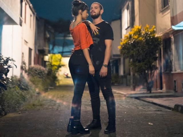 SpicyFran, con pechos pequeños, una blusa naranja, pantalones negros y botas negras, está de pie frente a un hombre con barba, de complexión musculosa, que lleva una camisa negra y pantalones negros, y que la está sosteniendo..