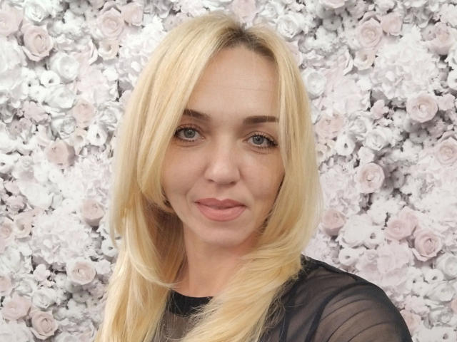 SallySweet-hot has blonde hair, blue eyes, and a cute smile. She's wearing a sheer black top, showing off her tits. Her pussy is not visible.