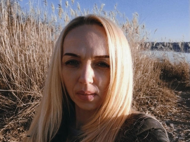 SallySweet-hot has blonde hair, a serious face, and is looking at the camera with slightly puffed-up lips. She's outdoors with tall grass.