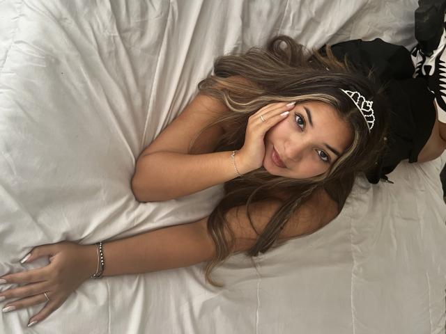 AmbarDunn with long brown hair, wearing a silver tiara and black lingerie, lying on white sheets, hand on face, looking up with a slight smile, showing her tits.
