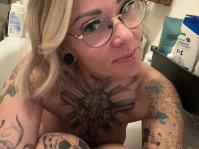 Lexi88-hot, topless in a bathtub, has tattoos on her chest and arms, wears glasses, nose ring, and black ear gauges, with blonde hair, smiling at the camera.