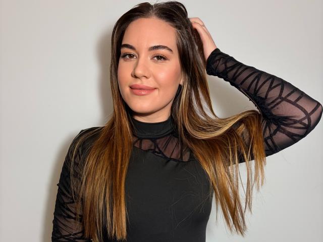 JosieDelicious with long brown hair, black sheer long-sleeve top, hand in hair, slight smile, light skin, and brown eyes, standing against a plain white background.
