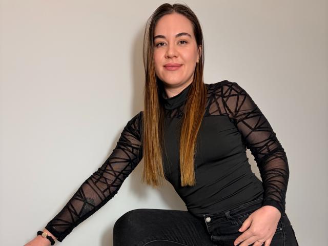 JosieDelicious has long brown hair, black sheer long-sleeve top, black jeans, and a slight smile, sitting with arms relaxed, showing a cute ass.