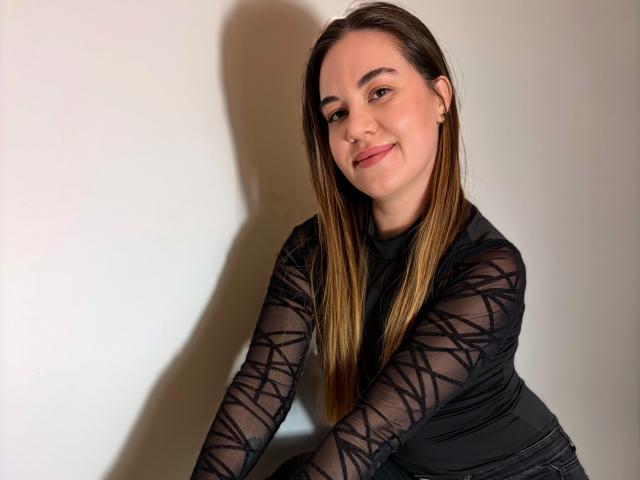 JosieDelicious with long brown hair, wearing a black sheer long-sleeve top with geometric patterns, smiling slightly, and showing a small nose ring.