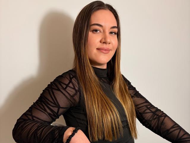 JosieDelicious has long brown hair, a cute face, and wears a black sheer top with geometric patterns, showing her tits through the fabric. She's smiling with hands on her hips.