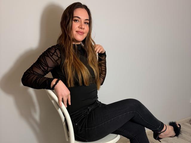 JosieDelicious with long brown hair, black sheer long-sleeve top, black jeans, and black furry slippers, sitting on white chair, hand on chair, other hand on chest.