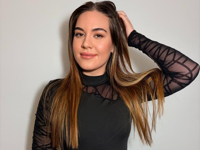 JosieDelicious with long brown hair, wearing a black sheer long-sleeve top, hand in hair, smiling softly, light skin, subtle makeup, medium-sized tits, casual confident pose.