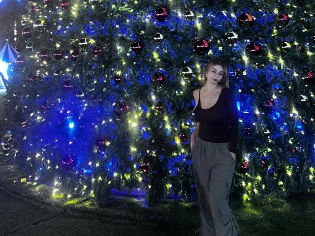 MarianneBowe stands in front of a Christmas tree, wearing a black top, gray pants, and smiling. Tree is adorned with red ornaments and blue lights.