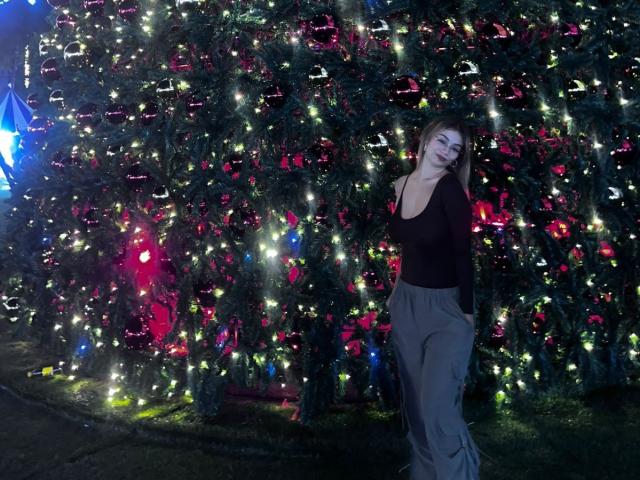MarianneBowe with long brown hair, black tank top, gray pants, standing in front of a Christmas tree with colorful lights and red ornaments, smiling slightly.