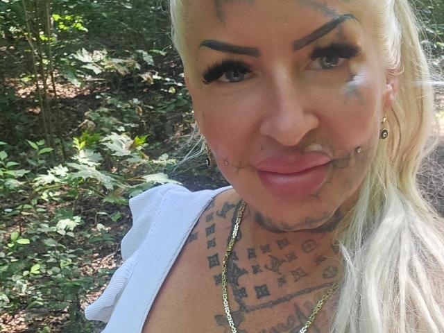 Jeanny-Bln, con il viso sporco di vernice blu e nera, capelli biondi, top bianco, tatuaggi sul collo, orecchini dorati e labbra rosa, in piedi in una foresta..
