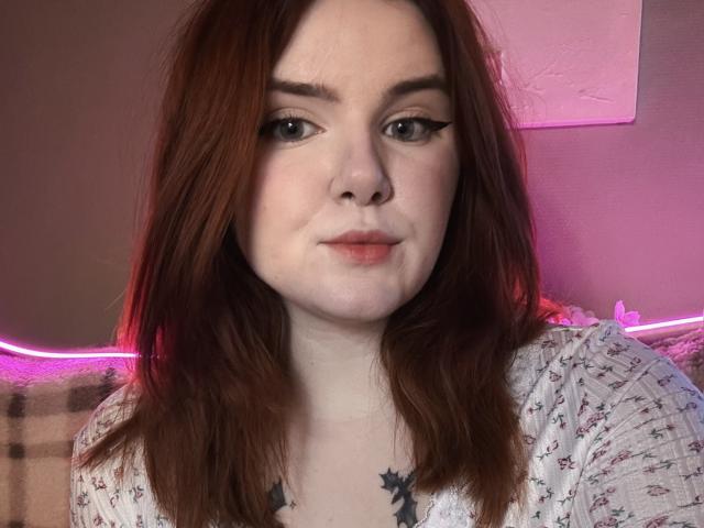 Close-up of Rubbi with fair skin and red-brown hair. She has black winged eyeliner, a small nose piercing, and a tattoo of a black bird on her chest. She wears a white, floral-patterned shirt. Soft pink and purple lighting in the background. Rubbi's expression is neutral, with slightly parted lips. The image has a casual, personal feel. Rubbi's hair is slightly messy. Her eyebrows are thick and dark.