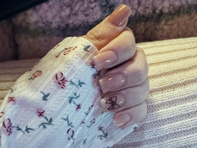Rubbi's hand holding another hand, both with glossy, nude-painted nails, wearing a white, floral-patterned, ribbed sleeve. Rubbi's fingers are slightly bent, showing a small, black butterfly nail art.