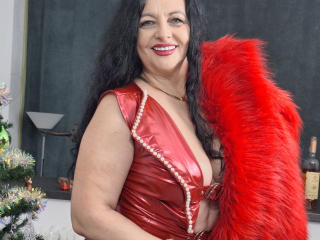 MelynnaBrown with long black hair, red shiny dress, and red feather boa, smiling at camera, showing her tits, standing near a decorated Christmas tree and bottle.