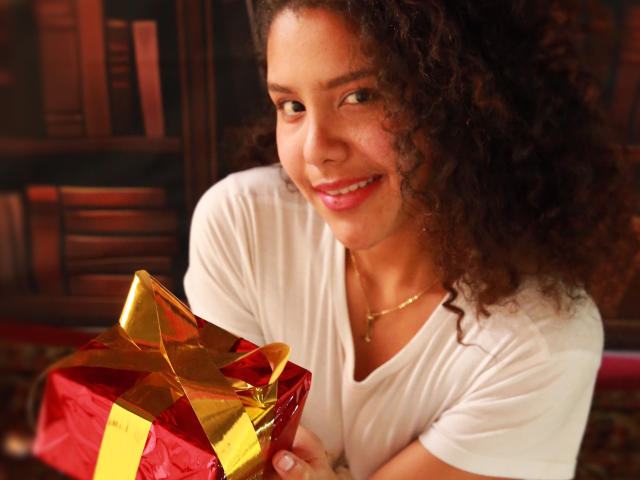 AriaVega with curly brown hair, smiling, holding a red and gold gift box, wearing a white shirt, in a cozy, warmly lit room. Her tits are small, cute.