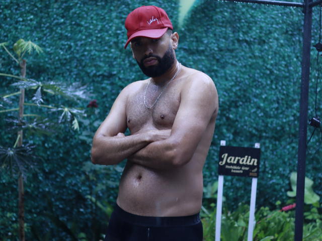 AntoniSmith è senza camicia, indossa un cappello rosso, pantaloni neri e una collana, ha la barba, le braccia incrociate e un petto leggermente muscoloso e peloso, ed è in piedi davanti a uno sfondo verde e pieno di foglie..