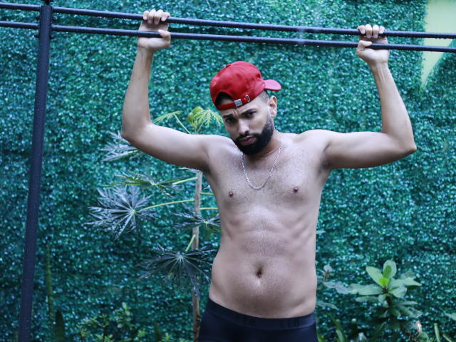 AntoniSmith est torse nu, il tient une barre les bras levés, il porte une casquette rouge et un short noir, il a une barbe, un collier en chaîne, et un léger ventre, et il regarde vers le bas..