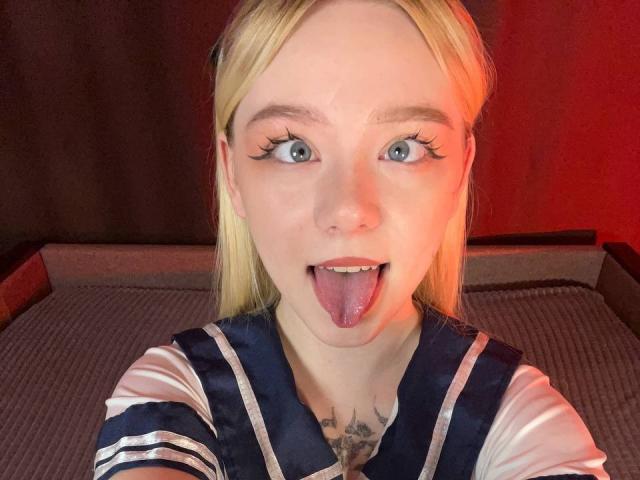 EnchantingEyes with blonde hair, green eyes, and a tattoo under the chin, sticking out her tongue, wearing a blue and white sailor-style top.