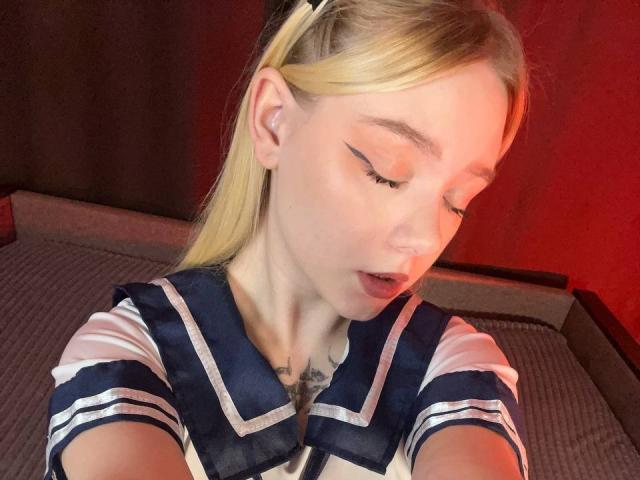 EnchantingEyes with blonde pigtails, eyeliner wings, and a navy sailor dress, showing off a chest tattoo, has her eyes closed and lips slightly parted.