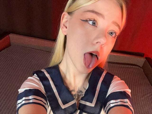 EnchantingEyes, blonde, with tongue out, wearing a navy and white sailor outfit, has a tattoo on her chest, and has sharp eyeliner, in a red-lit setting.